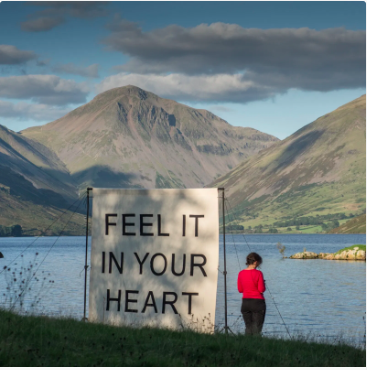 A canvas with the words Feel It In Your Heart erected beside a lake, with a woman standing next to it and mountains in the distance