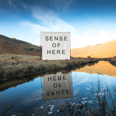 A canvas with the words Sense of Here on it placed next to a river and among hills, with a blue sky
